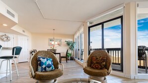 A spacious living area with two patterned armchairs, a glass coffee table with plants, a dining area with a chandelier, and large windows offering a view. - Sandpiper Condominiums 303L, Panoramic Gulf Views w/ direct access to Mustang Island/Port A beaches. (Port Aransas)
