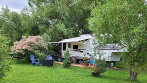 A recreational vehicle (RV) parked in a grassy area with a small deck, blue chairs, and a table.