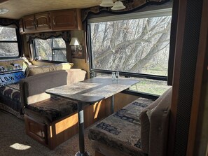 A cozy RV interior with a dining area, seating, and large windows offering a view of trees. - Unique Yellowstone Camping Glamping Getaway.   RV on Solo Sight w/ Full Hookups (Gardiner)