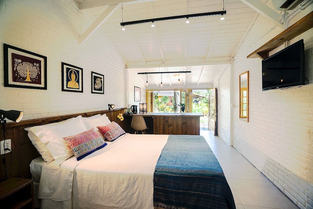 A bedroom with a large bed, a desk, and a TV. There are framed pictures on the wall and a window with a view of greenery.