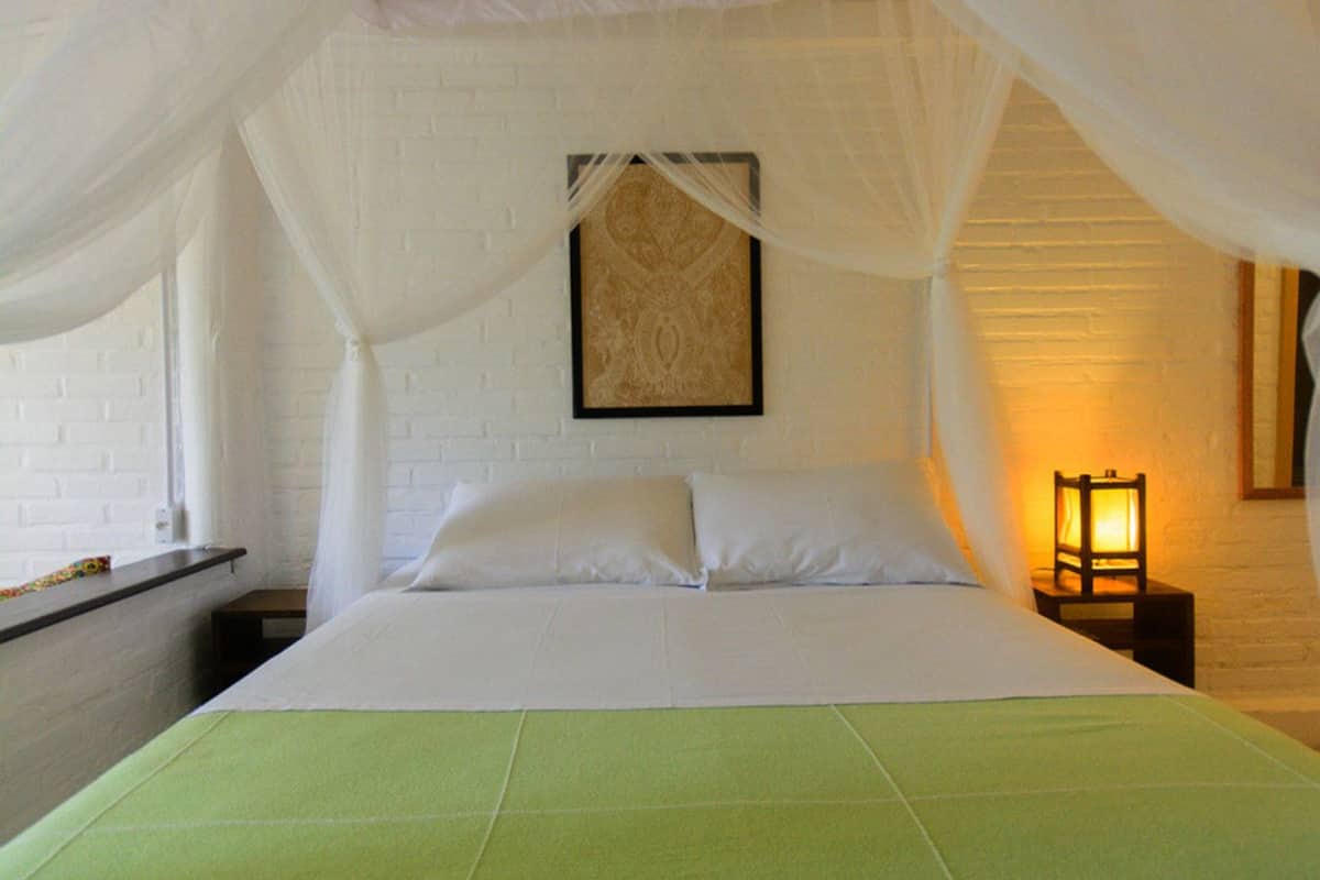 A bed with a green bedspread, white pillows, and a canopy. There is a framed picture on the wall and a small table with a lamp beside the bed.