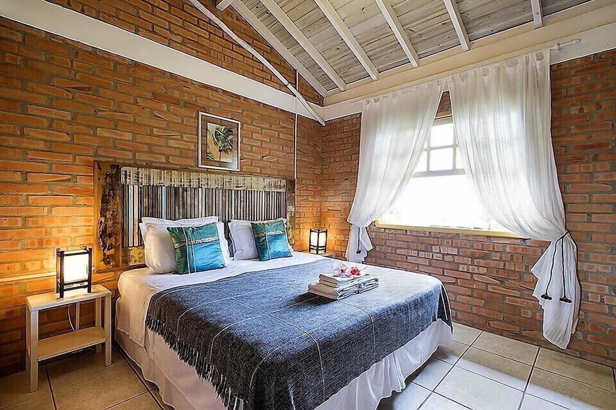 A bedroom with a brick wall, a bed with a blue bedspread, white pillows, and a headboard with vertical slats. There is a window with white curtains and a small table with a lamp beside the bed.