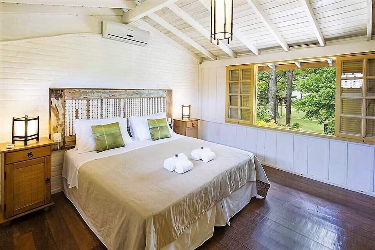 A bedroom with a large bed, wooden headboard, bedside tables with lamps, and a window with a view of greenery.