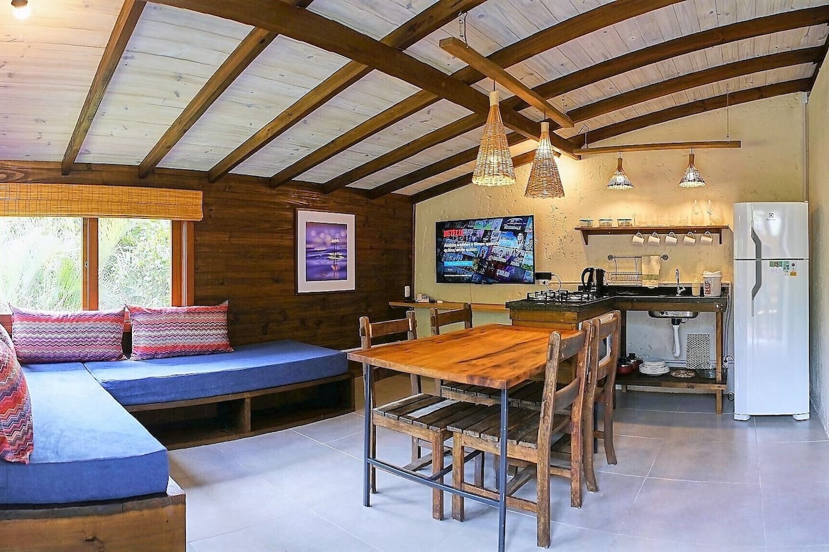 A cozy cabin interior with a wooden ceiling, a dining table with chairs, a kitchen area, and a built-in bench with cushions.