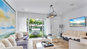 A modern living room with a sofa, coffee table, and a TV. There is a glass table with chairs outside, and a painting on the wall. -  Blissful Coral Villa EcoPool4b-4b (Miami)