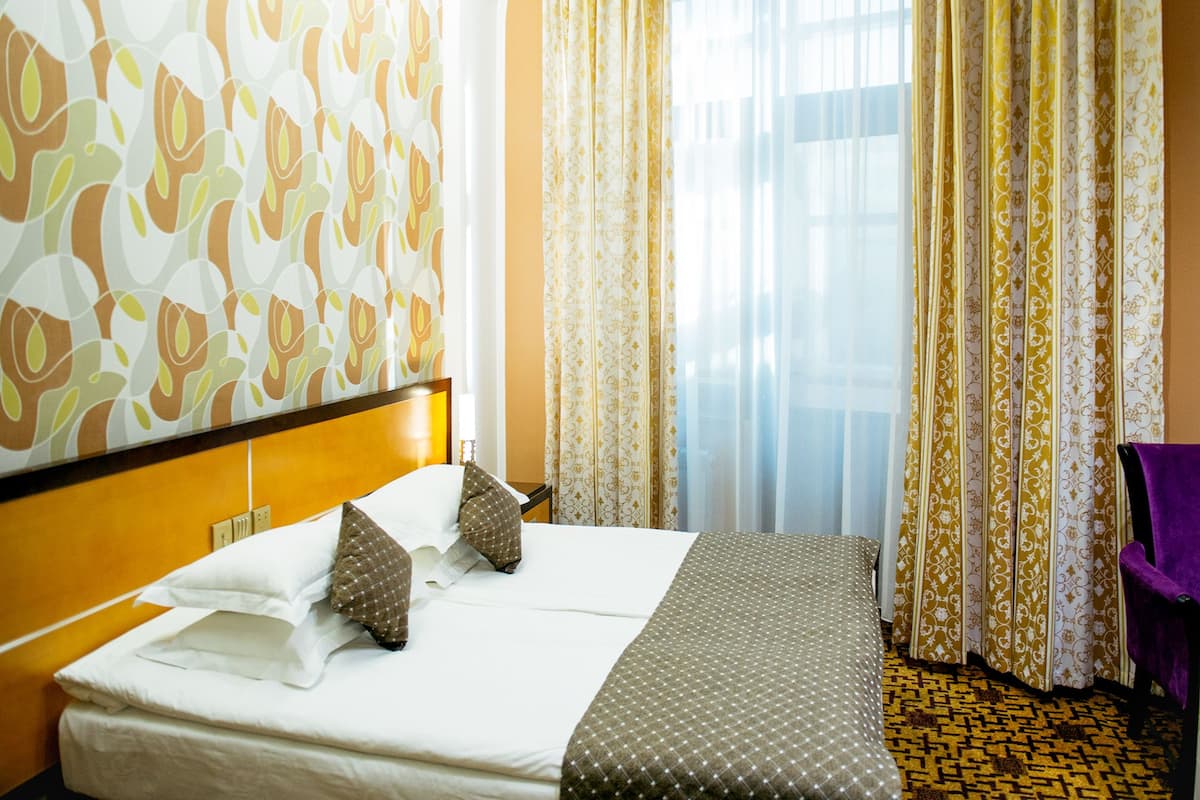 A hotel room with a large bed, a headboard, a window with curtains, and a patterned carpet.