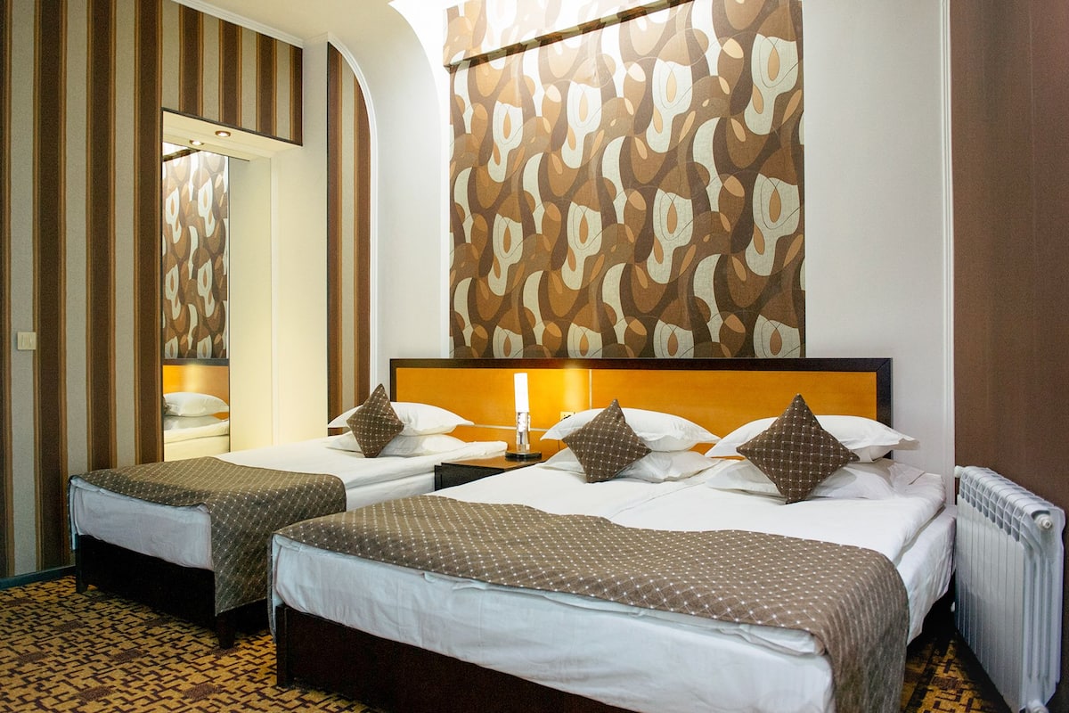 A hotel room with two single beds, a large mirror, patterned wallpaper, and a radiator.