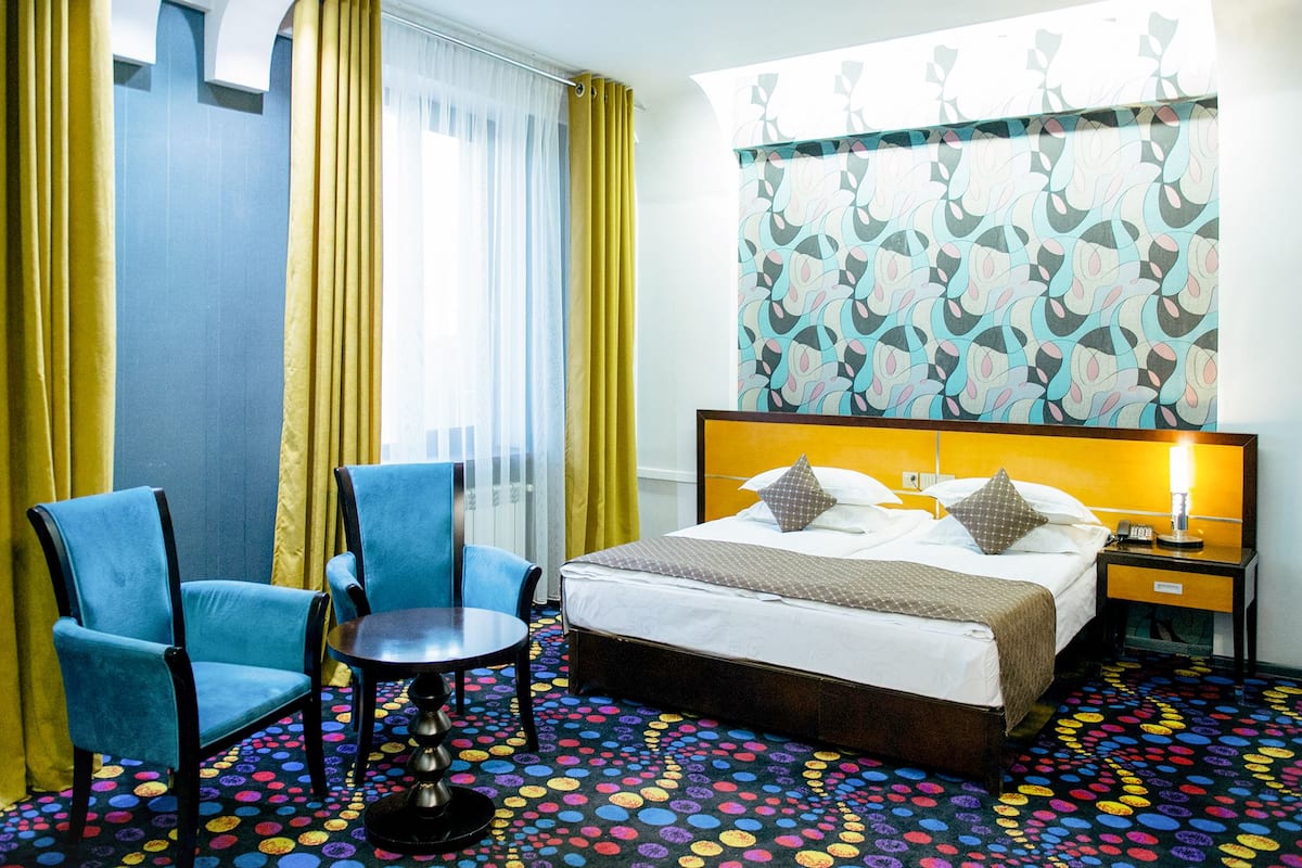 A hotel room with a bed, two blue chairs, a small table, a patterned carpet, and a window with curtains.