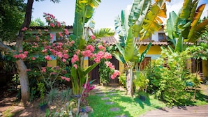 A garden with a wooden deck, tropical plants, and a yellow building with a tiled roof. - Pouso Sambaquis (Ilhabela)