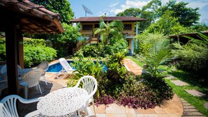 A pool area with lounge chairs and a table, surrounded by lush greenery and a house with a tiled roof. - Pouso Sambaquis (Ilhabela)