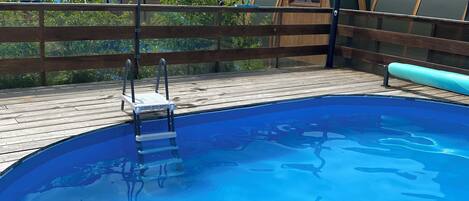 A swimming pool with a wooden deck, a ladder, and a covered area with a wooden door.