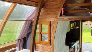 Interior acogedor de una cabaña con techo de madera, un comedor con mesa y sillas, una cocina con estufa y una ventana con vistas a un paisaje verde.