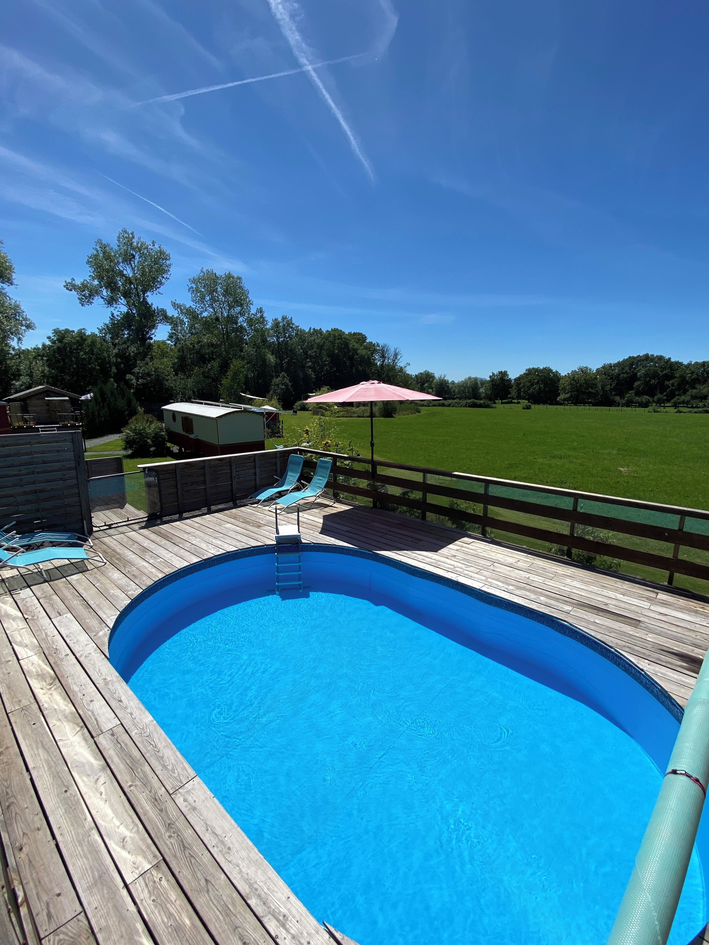 A wooden deck with a swimming pool, a pink umbrella, and a view of a green field and trees.