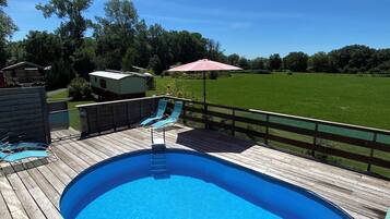 A wooden deck with a swimming pool, a pink umbrella, and a view of a green field and trees.