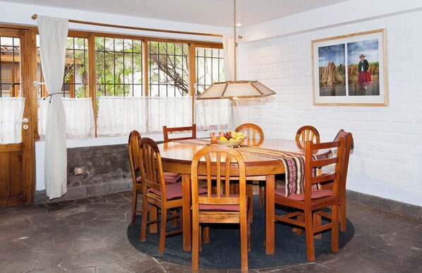Un comedor con una mesa redonda de madera, seis sillas de madera, una cesta de mimbre con fruta y un cuadro colgado en la pared.