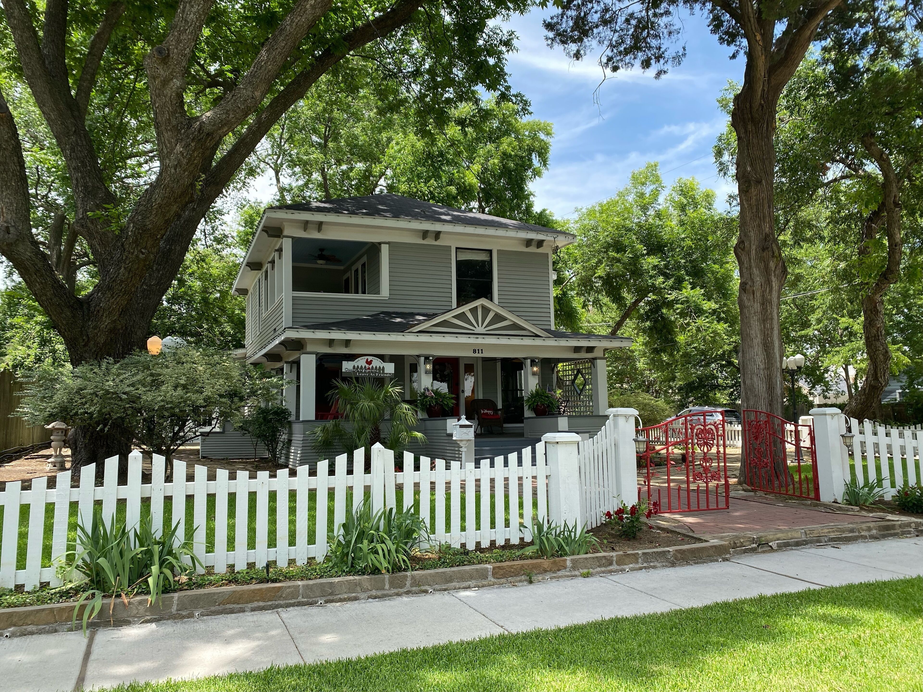 Une maison à deux étages avec un porche, entourée d’une clôture en piquets blancs et de grands arbres.