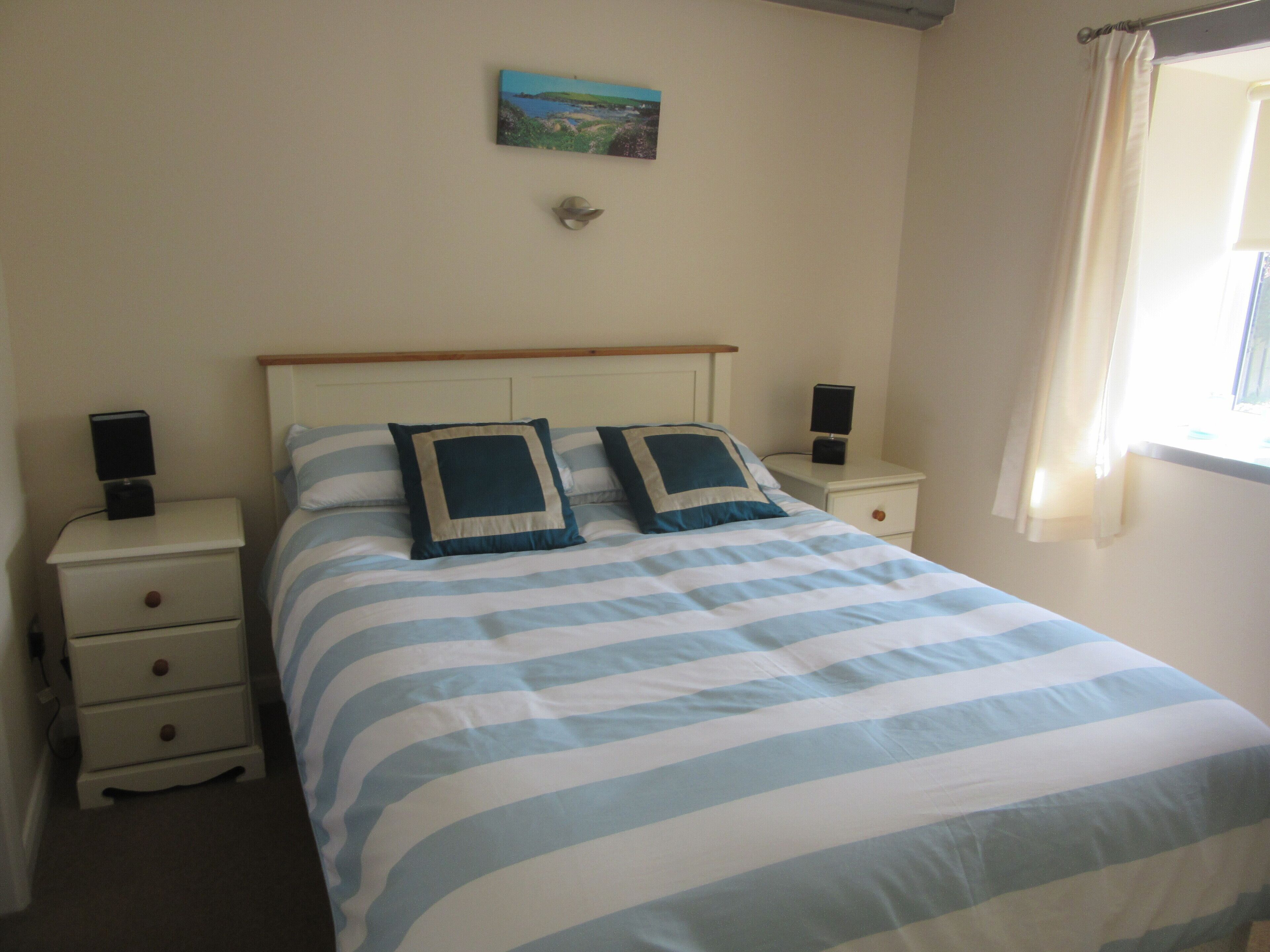 A bedroom with a bed, two bedside tables, a nightstand, a painting on the wall, and a window with curtains.