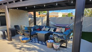 An outdoor patio with a covered seating area, including a sofa and chairs, surrounded by potted plants and a patterned rug. - Spacious Mesa Oasis with FREE Heated POOL (Mesa)