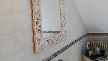 A bathroom with a white tiled wall, a decorative mirror with an ornate frame, a wall-mounted lamp, and a small shelf with toiletries.