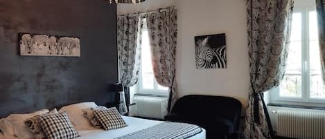 Un dormitorio con cama, una silla negra, una araña, cortinas estampadas y obras de arte en las paredes.