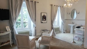 A room with a bed, two wicker chairs, a vanity with a mirror, a radiator, a television, and a chandelier. - Chateau les 4 saisons (Saint-Cirgues-sur-Couze)