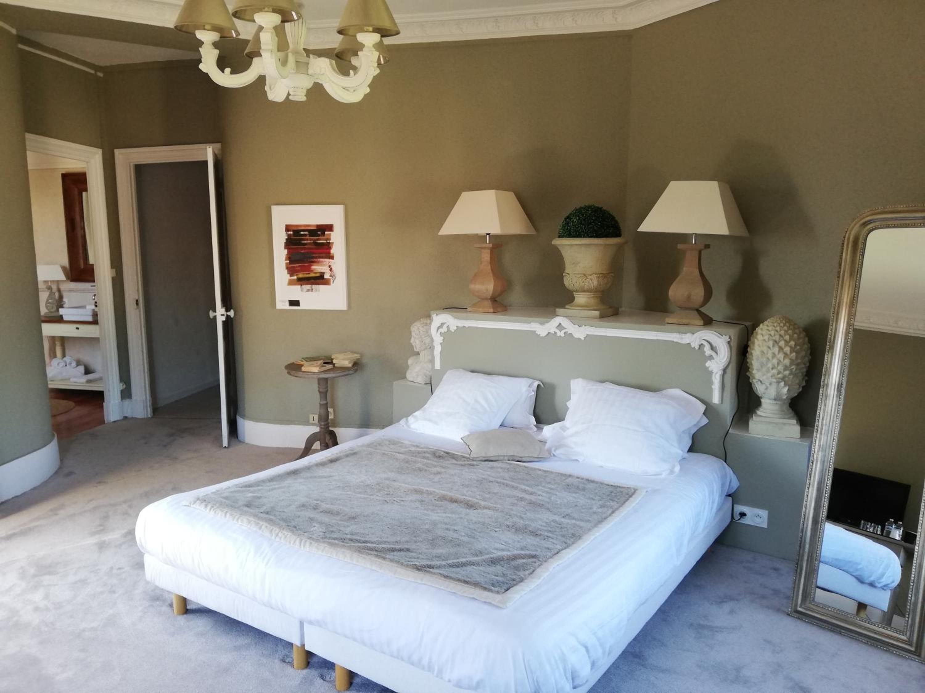 Ein Schlafzimmer mit einem großen Bett, zwei Nachttischlampen, einem kleinen Tisch, einem Spiegel und einer Tür zu einem anderen Raum.