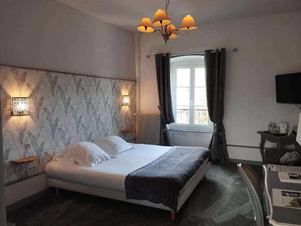 A hotel room with a large bed, a desk with a chair, a window with curtains, and a chandelier.