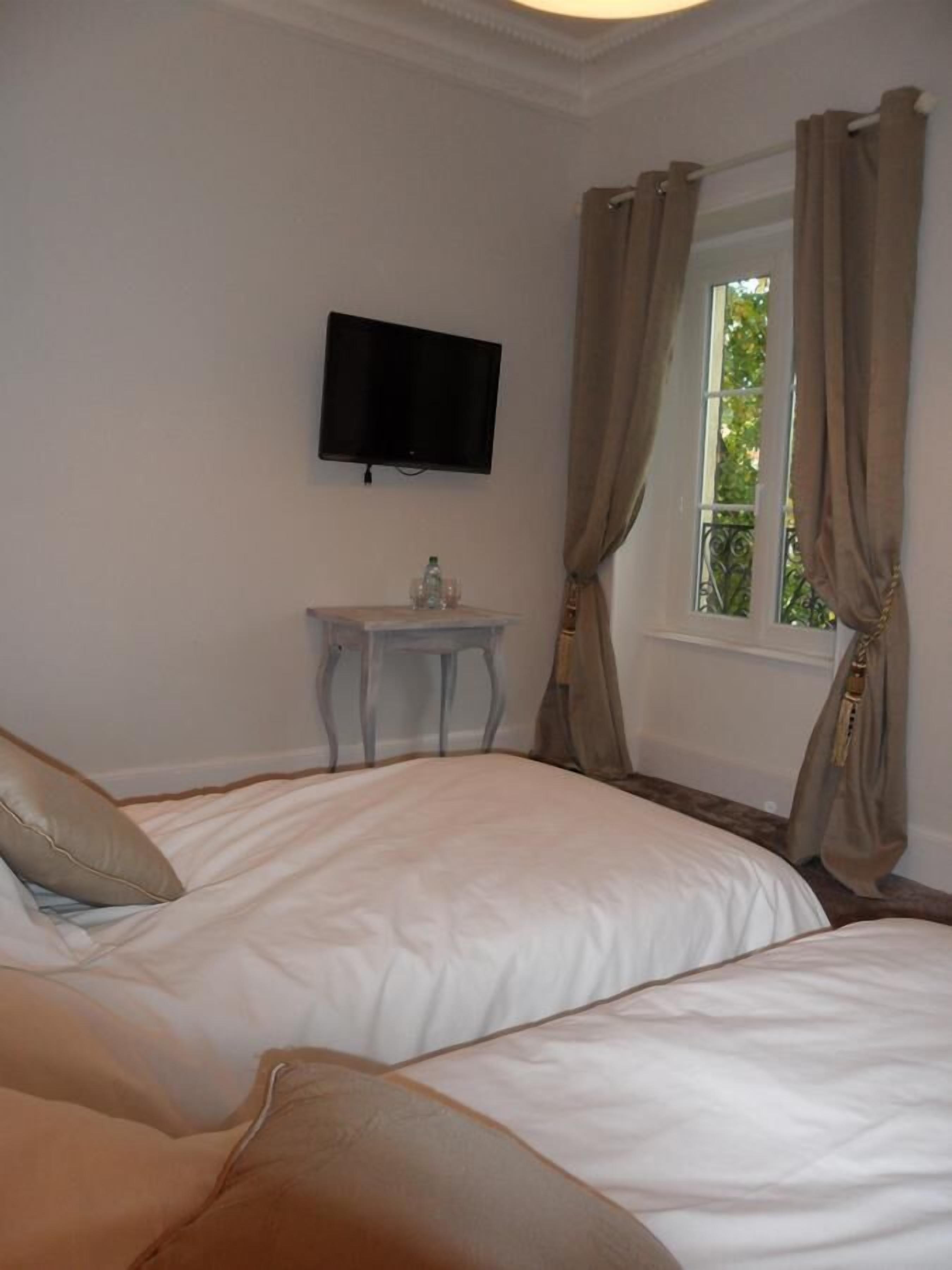 A hotel room with two beds, a small table, a TV mounted on the wall, and a window with curtains.