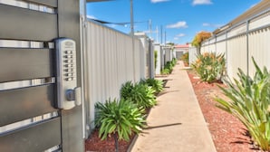 A pathway flanked by metal fences and small plants, leading to a building with a keypad lock.