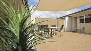 An outdoor patio area with a table and chairs, a large shade sail, and a building in the background.