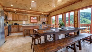 A spacious kitchen with wooden cabinets and a large island, a dining area with benches, and a view of a forested landscape through large windows. - Private Heated Indoor Pool, Mountain View, Hot Tub, Pigeon Forge Cabin, Pool Table, Theater Room (Sevierville)