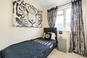 A bedroom with a bed, a nightstand, a lamp, a window with curtains, and a tiger painting on the wall. - Weybridge House (Weybridge)