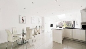 Una cocina moderna con armarios blancos, una mesa de comedor de vidrio y sillas. Hay cuadros en la pared y un ventanal que proporciona luz natural.