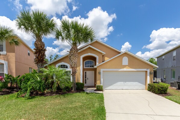 A two-story house with a beige exterior, a white garage door, and a well-maintained lawn with palm trees. - Minnies Emerald by VillaDirect (Kissimmee)