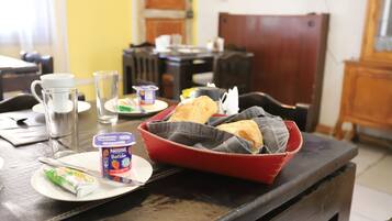 A breakfast table with a red basket containing bread rolls, a plate with a Nestle Batide yogurt, a glass of water, and a packet of tea.