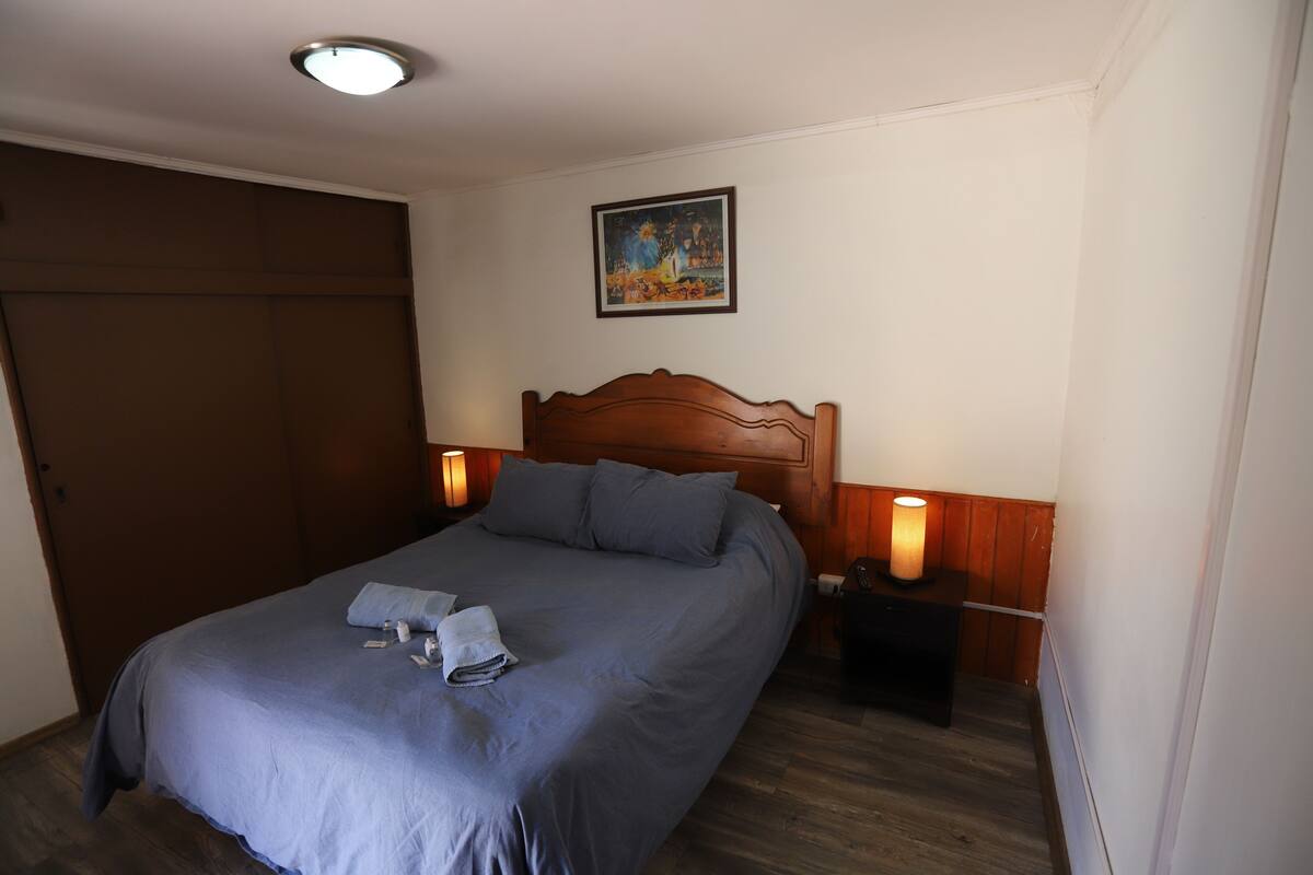 Una cama bien hecha con cabecera de madera, una mesita de noche con lámpara y un cuadro en la pared.