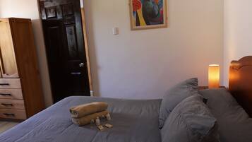 Un dormitorio con una cama, almohadas, una lámpara de noche, un armario de madera, una puerta y un cuadro en la pared.