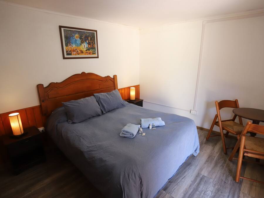 Un dormitorio con una cama grande, cabecera de madera, dos mesitas de noche con lámparas, una pequeña mesa redonda con sillas y un cuadro en la pared.