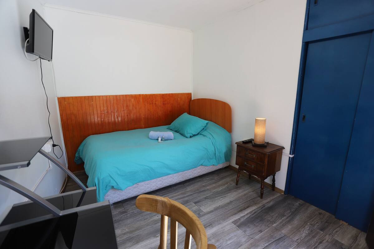 Un dormitorio con una cama, un escritorio, una silla, una mesita de noche con una lámpara y una puerta azul.