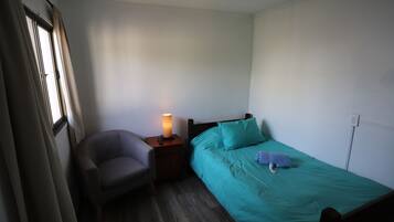 A small, single-bed room with a wooden nightstand, a lamp, a chair, and a window with curtains.