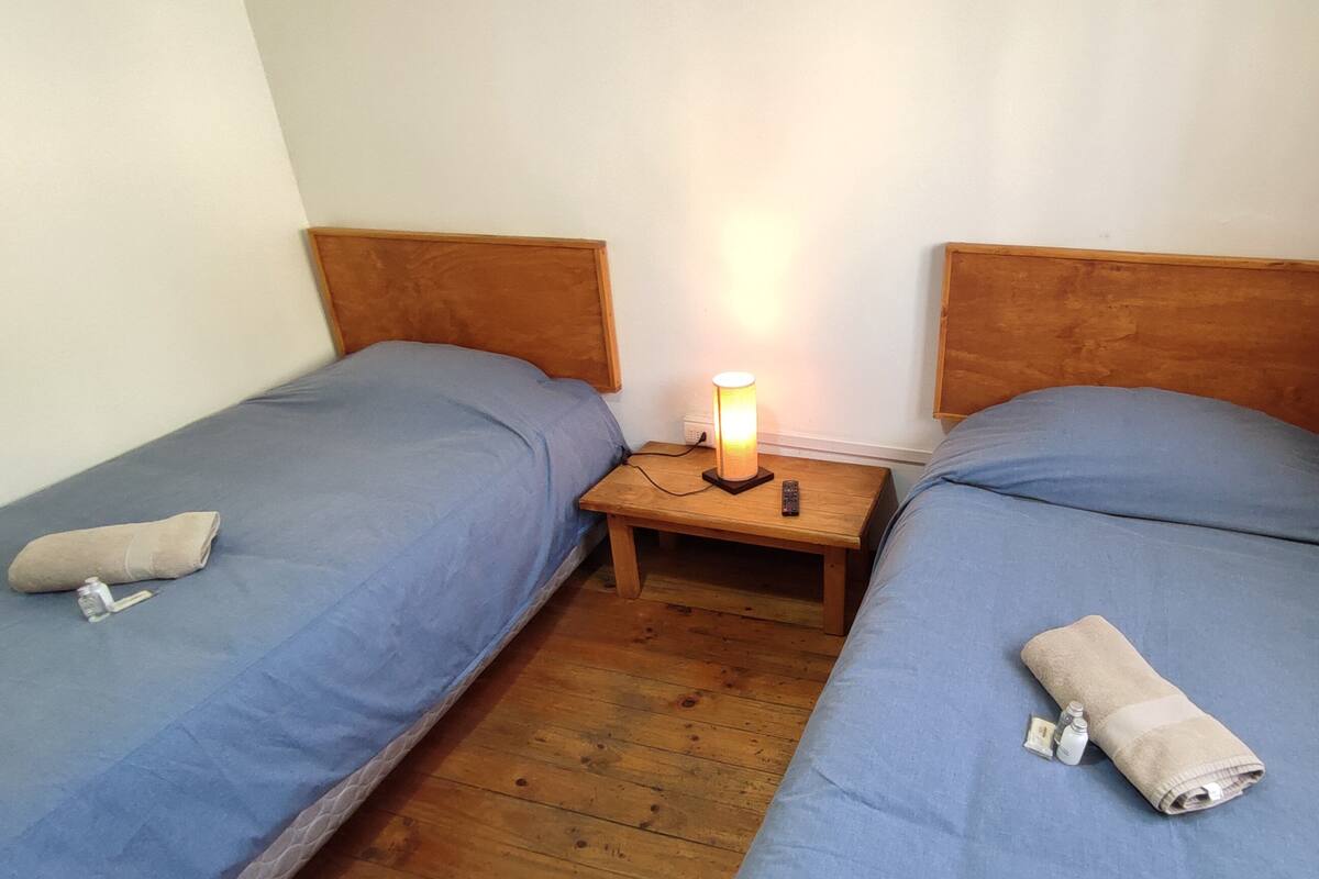 Una habitación con dos camas individuales, cada una con cabecero de madera, una mesita de noche con una lámpara y una toalla enrollada sobre cada cama.