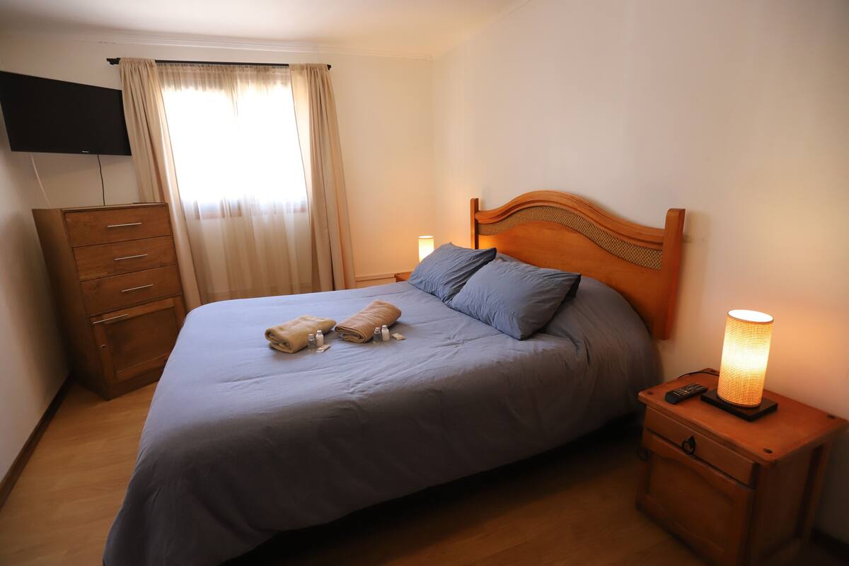 Una cama bien hecha con un cabecero de madera, una lámpara de noche y una mesita de noche de madera con un control remoto.