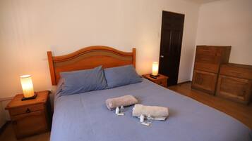 Een opgemaakt bed met blauw beddengoed, twee nachtkastjes met lampen, een houten garderobe en een deur.