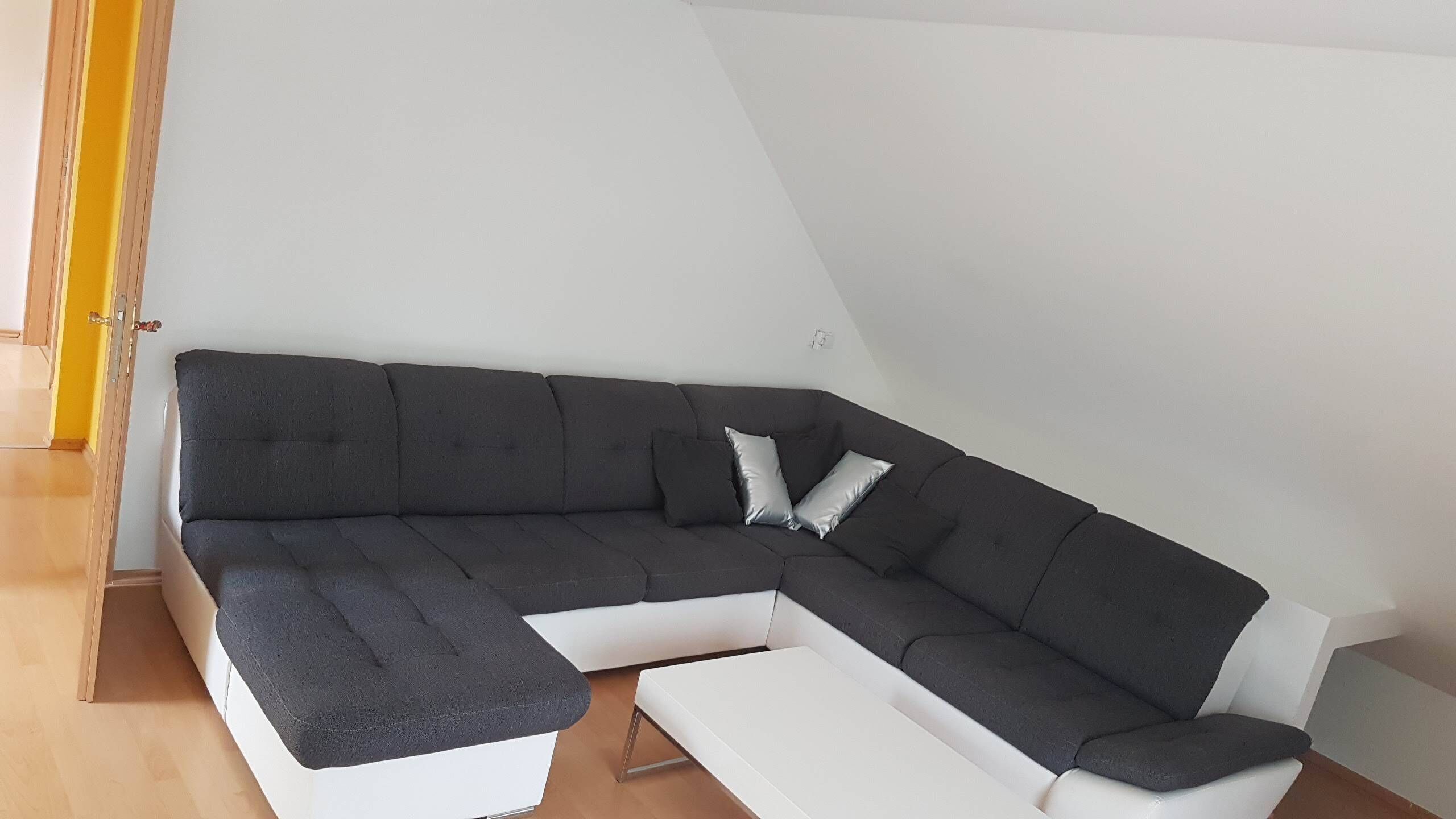 Un sofá modular moderno en forma de L con un detalle blanco, ubicado en una habitación con piso de madera y paredes blancas.