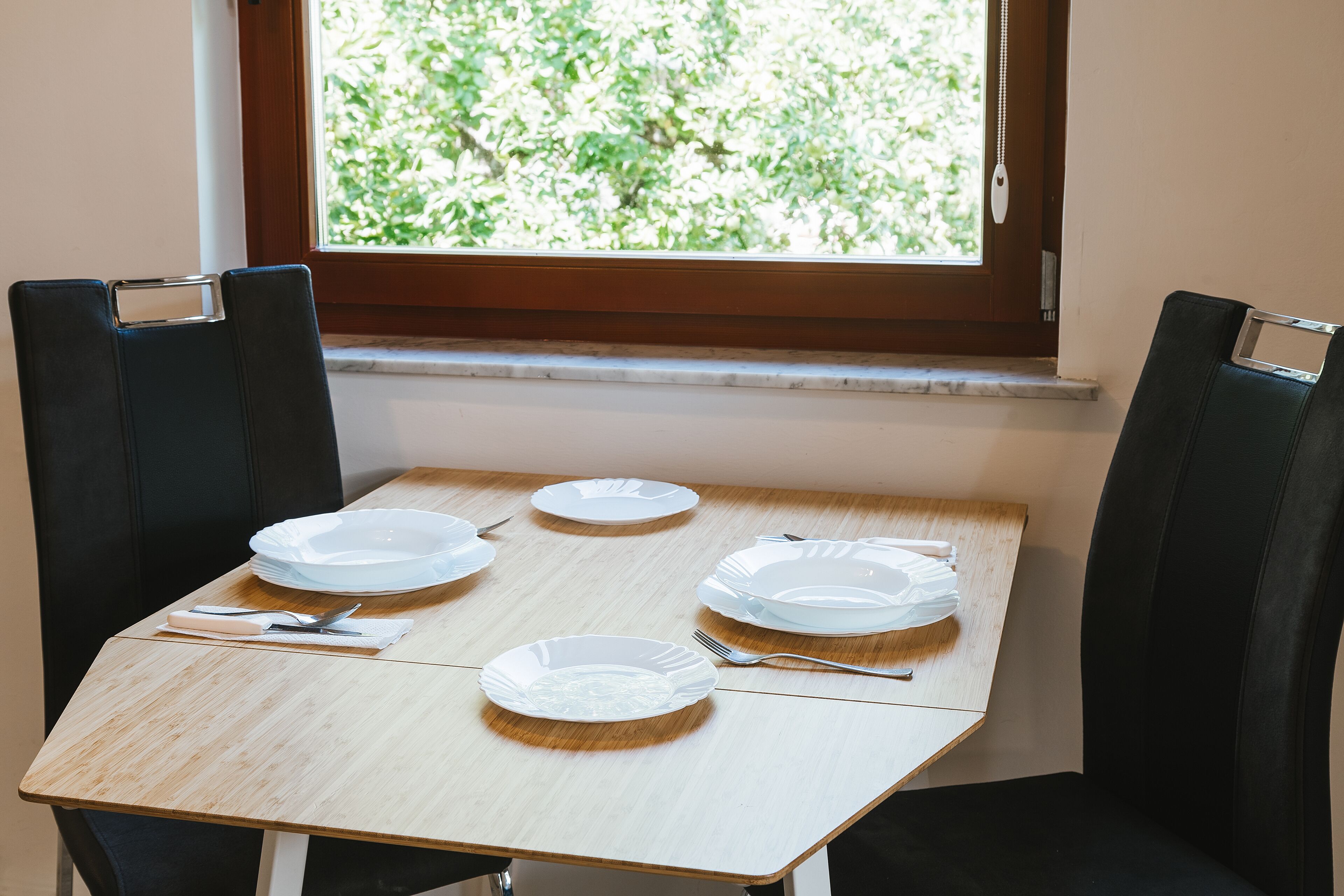 Une table à manger avec quatre assiettes blanches, des couverts et une serviette. La table est placée devant une fenêtre donnant sur un paysage verdoyant.
