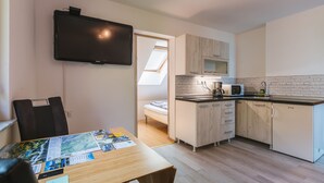 Une cuisine compacte avec un téléviseur à écran plat, une table à manger recouverte de papiers et un petit lit à l’arrière-plan.