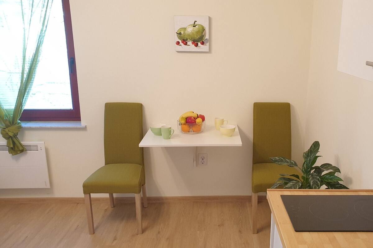 Una habitación pequeña y luminosa con dos sillas verdes, una mesa blanca con un frutero y tazas, una planta en maceta y una ventana con cortinas.