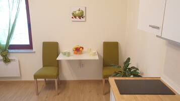 Una habitación pequeña y luminosa con dos sillas verdes, una mesa blanca con un frutero y tazas, una planta en maceta y una ventana con cortinas.