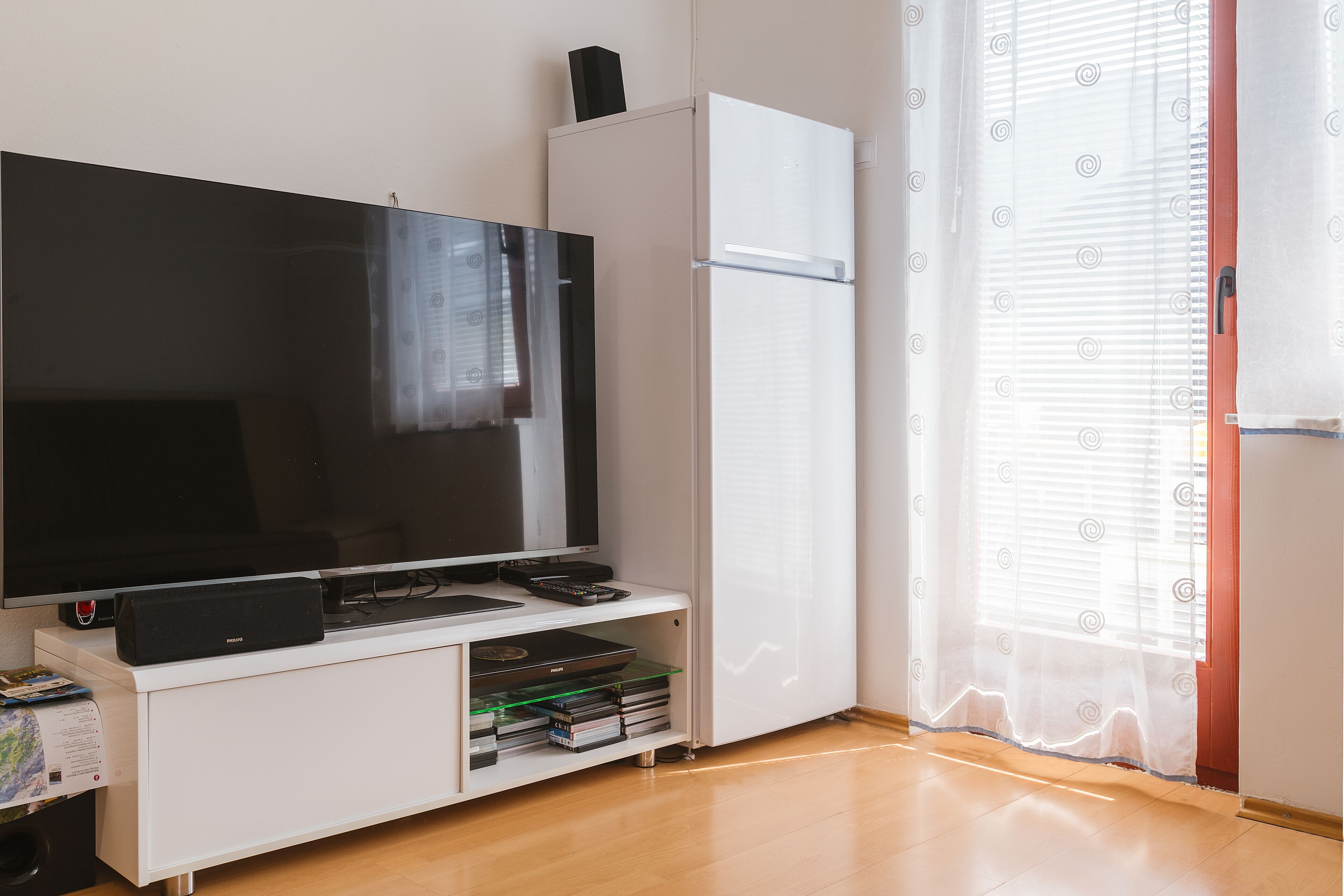 A modern living room with a flat-screen TV, a white cabinet, and a glass shelf with books and electronic devices.