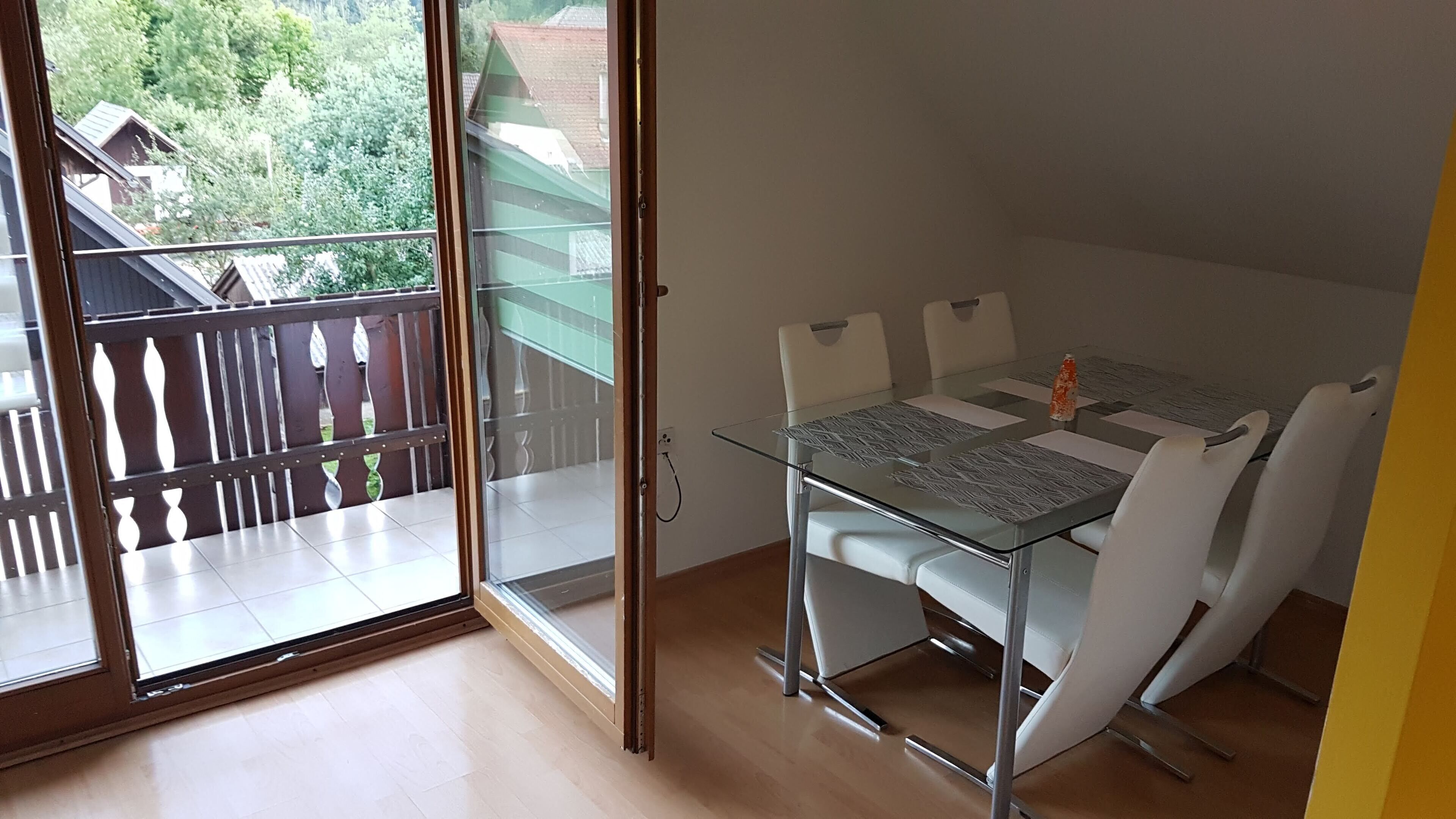A small balcony with a glass door, a glass table with white chairs, and a view of greenery and buildings outside.
